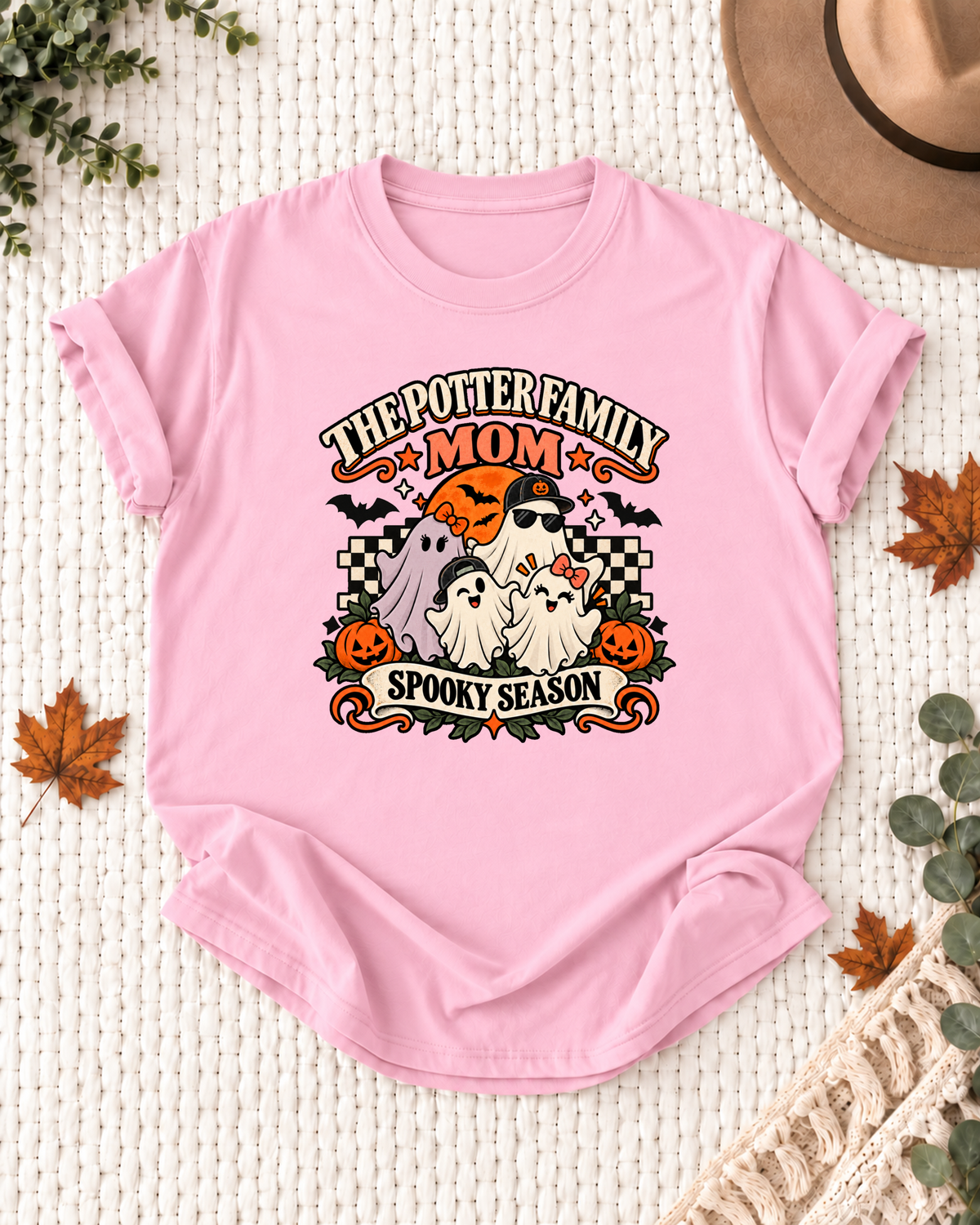 Light pink Mom T-shirt with four friendly ghosts, jack-o'-lanterns, bats, an orange moon, black-and-cream checkerboard accents, and example family-name text.