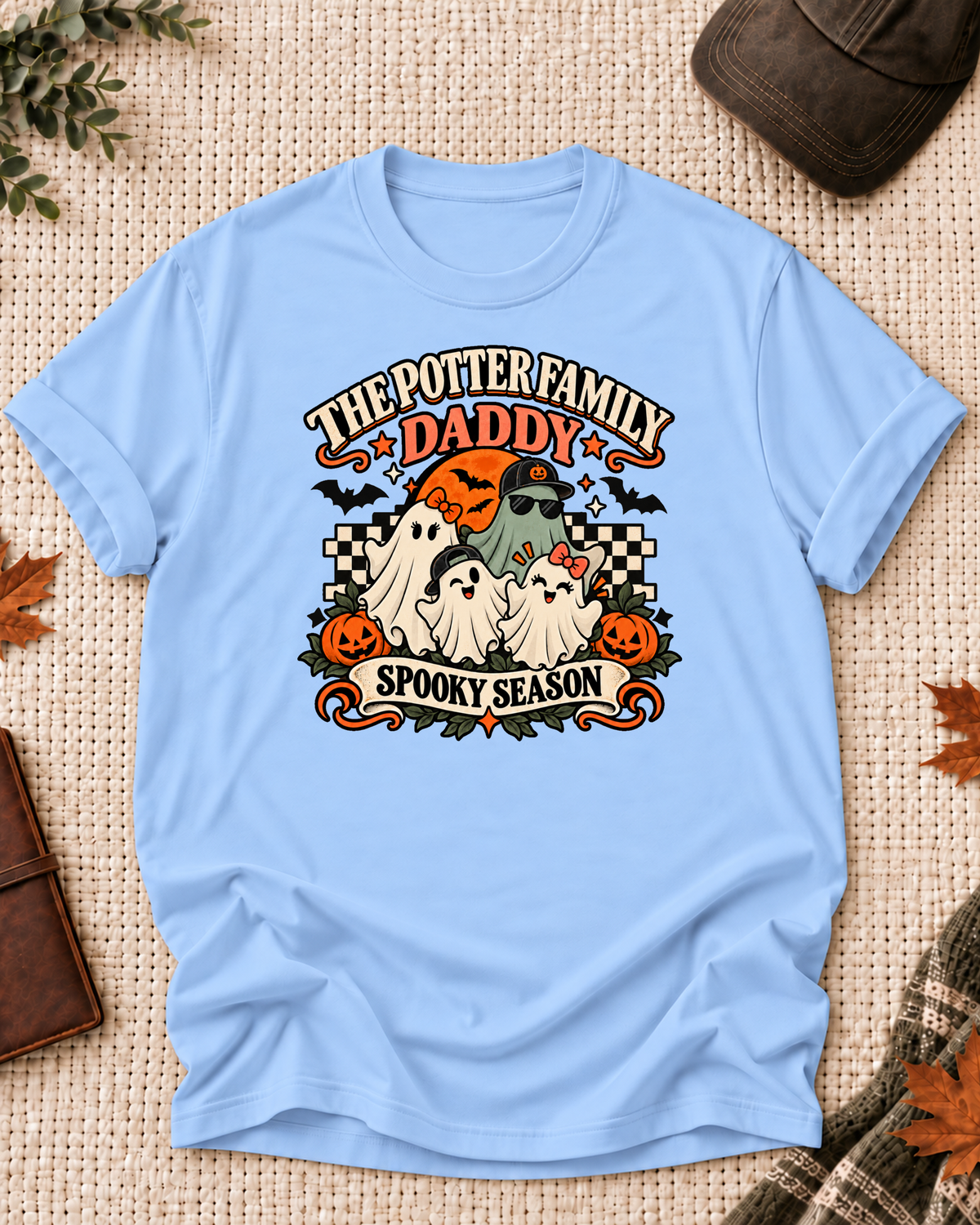 Light blue Daddy T-shirt with four friendly ghosts, jack-o'-lanterns, bats, an orange moon, black-and-cream checkerboard accents, and example family-name text.
