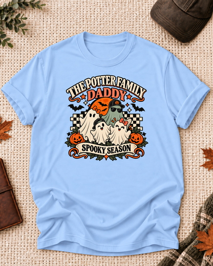 Light blue Daddy T-shirt with four friendly ghosts, jack-o'-lanterns, bats, an orange moon, black-and-cream checkerboard accents, and example family-name text.