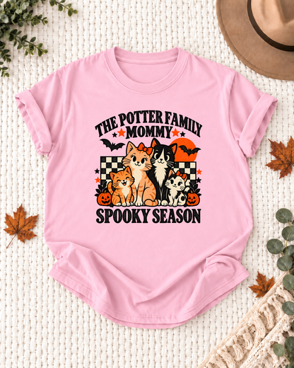 Light pink Mommy T-shirt with four illustrated cats, an orange moon, bats, black-and-cream checkerboard accents, jack-o'-lanterns, and example family-name text.