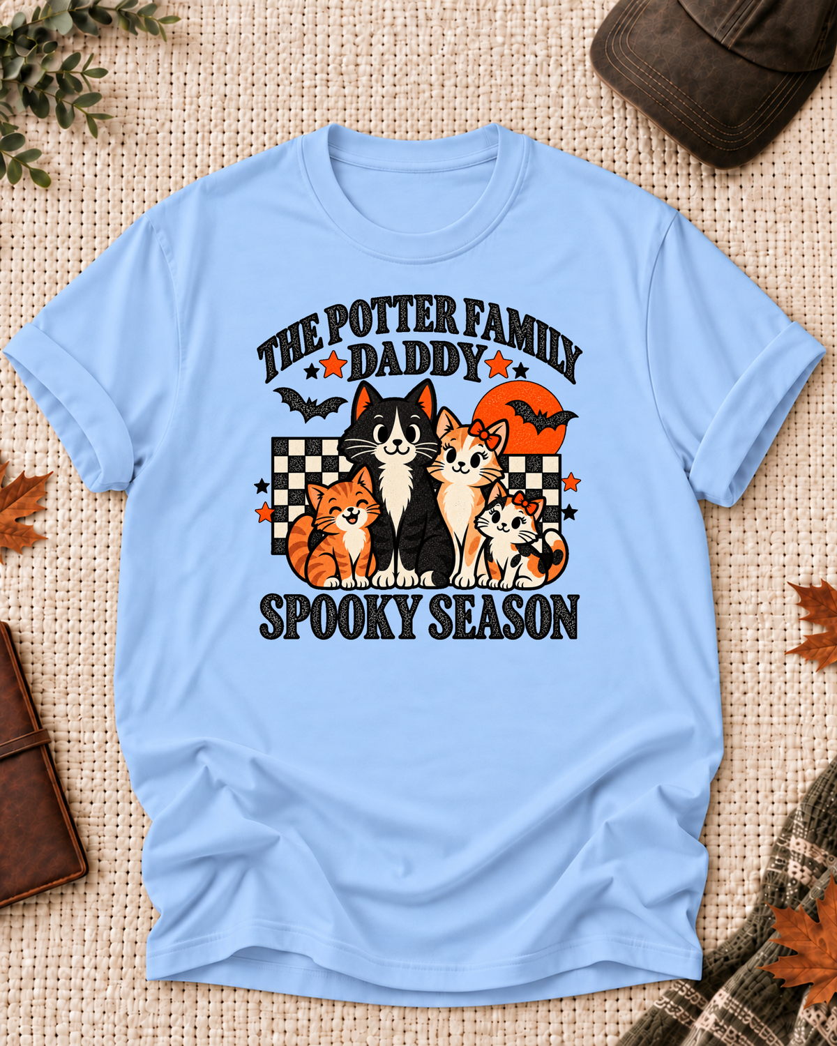 Light blue Daddy T-shirt with four illustrated cats, an orange moon, bats, black-and-cream checkerboard accents, jack-o'-lanterns, and example family-name text.