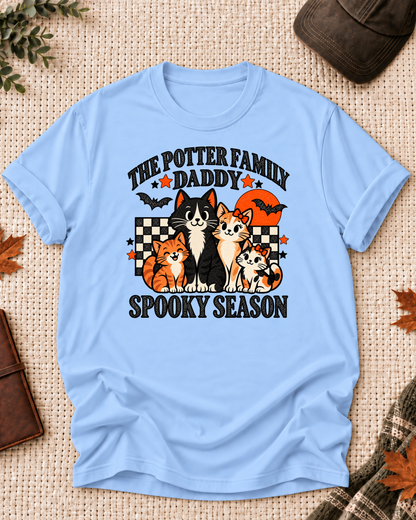 Light blue Daddy T-shirt with four illustrated cats, an orange moon, bats, black-and-cream checkerboard accents, jack-o'-lanterns, and example family-name text.