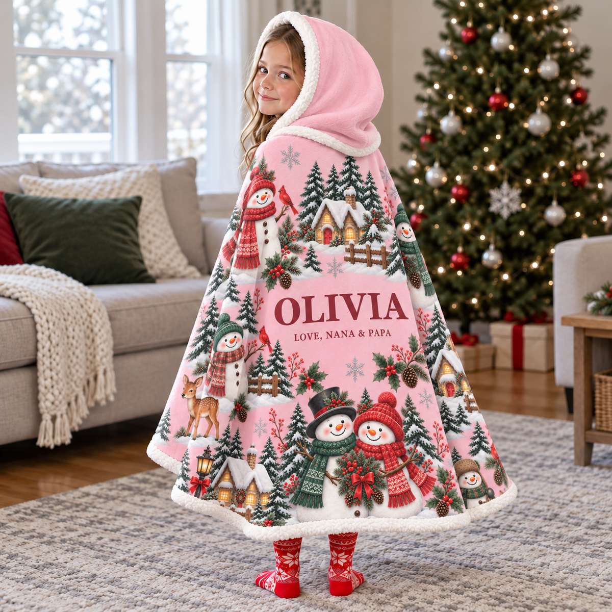 Child wearing a pink snowman hooded blanket by a Christmas tree, with example name Olivia and signature Love, Nana & Papa.