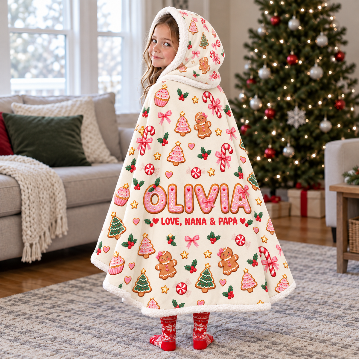 Child wearing an ivory gingerbread Christmas hooded blanket by a Christmas tree, with example name Olivia in pink and red signature Love, Nana & Papa.