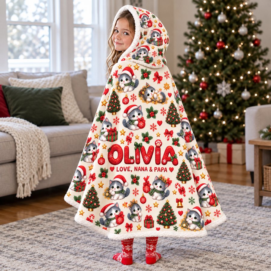 Child wearing an ivory Christmas kitten hooded blanket by a Christmas tree, with red example name Olivia and signature Love, Nana & Papa.