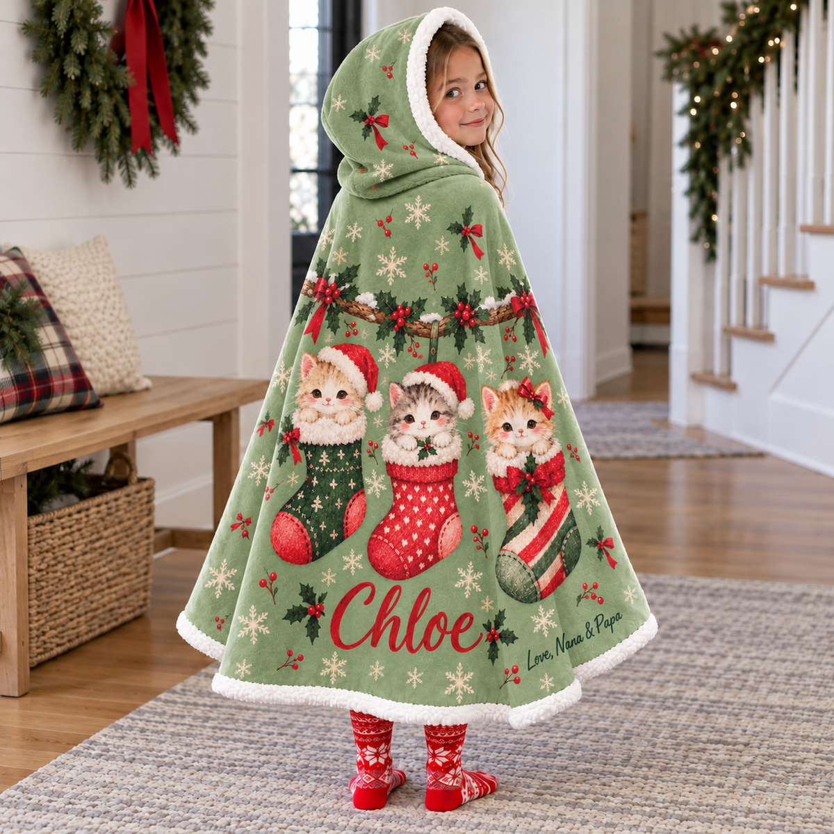 Child wearing a sage-green hooded blanket with three kittens in Christmas stockings, red example name Chloe, and dark green signature Love, Nana & Papa.